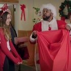 Menschen in festlicher Kleidung halten Geschenke. Ein Mann in einem Weihnachtsmannkostüm spricht. Weihnachtsschmuck im Hintergrund.
