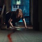 Keine sichtbaren Texte. Eine Frau wischt kniend den Boden, eine rote Flüssigkeit führt nach draußen, es regnet.