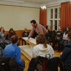 Personengruppe in Stuhlkreis, jemand zeigt etwas auf einem Tisch. Mehrere tragen Pullover mit Städtenamen drauf.