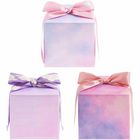 Drei würfelförmige Geschenke mit pastellfarbenem Papier und Schleifen, zwei rosa, ein lila mit Wolkenmuster.
