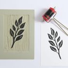 Ein Druck von Blättern, daneben eine Farbrolle und die Vorlage mit geschnitztem Blattmotiv auf Linoleum.
