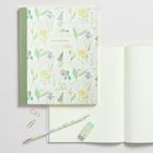„NOTEBOOK“ steht auf einem floralen Umschlag, daneben ein offenes Notizbuch, Bleistift, Radiergummi und Büroklammern.