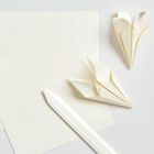 Blatt Papier, ein Faltwerkzeug und zwei gefaltete Origami-Herzen, alles in Weißtönen auf einem weißen Hintergrund.
