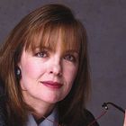 Es gibt keine Texte zu erwähnen. Eine Frau mit braunem Haar hält eine Brille, trägt einen Anzug und lächelt leicht.