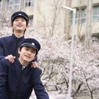 Zwei Personen in dunklen Uniformen lachen, im Hintergrund blühende Kirschbäume und ein Gebäude.
