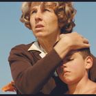 Eine Frau umarmt einen Jungen vor klarem blauem Himmel. Sie wirkt beschützend und nachdenklich.