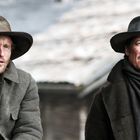 Zwei Männer in rustikaler Kleidung und Filzhüten sitzen nebeneinander, der Hintergrund ist unscharf.