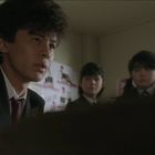 Ein junger Mann in einem Anzug schaut ernst, umgeben von mehreren Menschen in einem Raum mit Plakaten an der Wand.