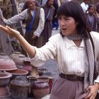 Eine Frau gestikuliert lebhaft mit ausgestrecktem Arm. Im Hintergrund stehen viele Menschen. Vor ihr Tongefäße.