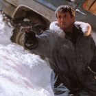 Ein Mann in Winterkleidung hält ein Gewehr in einer verschneiten Umgebung neben einem Fahrzeug.