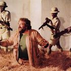 Ein Mann kniet in Stroh, umringt von vier bewaffneten Soldaten in Safari-Uniformen mit Helmen.