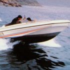 Seitlich "SD518". Zwei Personen in einem Motorboot, fahren schnell auf einer Wasseroberfläche. Küste im Hintergrund.