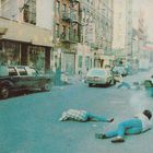 Texte: "Marcelo" und "Coca". Menschen liegen auf einer Straße, Autos parken daneben, städtische Szene.