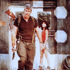 Ein großer Mann mit Gewehr in Tarnkleidung, eine Frau im Hintergrund, eine rustikale Umgebung mit großen Zahl "8" an der Wand.