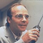 Ein Mann mit Brille und Anzug hält ein Handfunkgerät und lächelt.