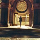 In einem großen Raum mit dekorativen Säulen und Mustern tritt eine Person in dunkler Kleidung durch Türen in warmes Licht.