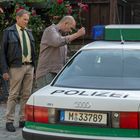 Ein Polizeiauto mit der Aufschrift "POLIZEI". Zwei Männer stehen daneben, einer durchsucht den Kofferraum. Im Hintergrund blühende Rosen.