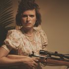 Keine sichtbaren Texte. Frau mit lockigem Haar trägt eine Blumenkleid und Perlenkette, hält Gewehr, wirkt entschlossen.