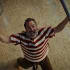 Ein Mann mit gestreiftem Pullover hebt freudig einen riesigen Büroklammer-Nachbau und einen Schläger in die Luft.