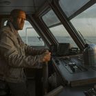 Ein Mann in einer Schiffssteuerkabine, konzentriert auf das Fahren bei Sonnenuntergang, umgeben von Kontrollinstrumenten.