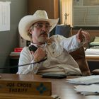 Ein Schild mit der Aufschrift "Joe Cross, Sheriff" liegt auf einem Schreibtisch. Ein Mann mit Hut telefoniert und zeigt.