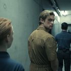 In einem kargen Flur gehen Personen; eine Person schaut über die Schulter zurück. Die Beleuchtung ist gedämpft.