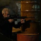 Text: "46LB". Ein älterer Mann mit Gewehr, im Schatten, hinter einem rostigen Fass in einer nächtlichen Szene.