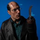 Eine Person in einer Lederjacke hat eine Glatze mit Narbe und trägt eine Brille. Ihr Arm endet in einem Haken.