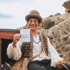 Ein Mann in historischer Kleidung hält ein Schild mit „GRAND RE-BIG-OPENING“. Er sitzt auf einer roten Kutsche vor Felsen.