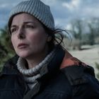 Eine Frau in Winterkleidung schaut ernst in die Ferne, mit Nadelbäumen und bedecktem Himmel im Hintergrund.