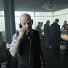 Ein Mann mit Glatze in einem Büro spricht in einen schwarzen Telefonhörer. Mehrere Personen im Hintergrund.