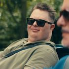 Zwei Männer mit Sonnenbrillen sitzen in einem Auto. Der Mann links trägt ein grünes Polohemd und lächelt leicht.