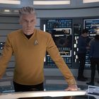 Ein Mann in gelber Uniform mit futuristischem Abzeichen steht ernst an einem Tisch in einem Raum mit Bildschirmen.