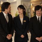 Drei Personen in eleganten Anzügen stehen nebeneinander. Sie tragen Namensschilder und stehen in einem dekorierten Raum.