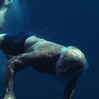 Kein sichtbarer Text. Ein älterer Mann taucht in klarem, blauem Wasser, trägt eine dunkle Badehose.