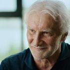 Ich kann die Texte im Bild nicht erkennen. Ein älterer Mann mit weißen Haaren schaut lächelnd zur Seite.