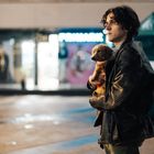 Ein junger Mann steht draußen, hält einen kleinen Hund im Arm, im Hintergrund verschwommene Lichter einer Einkaufspassage.