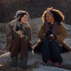 Zwei Personen sitzen draußen auf Steinen, warm gekleidet in Mäntel und Mützen, essen und trinken, umgeben von trockenem Gras.