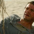 Ein Mann liegt unter Palmenblättern am Strand, blutverschmiert und besorgt. Er trägt eine Uhr und ein blaues Hemd.