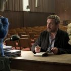 Ein Mann signiert ein Buch am Tisch; Orchidee und Bücherstapel daneben. Frau im blauen Pullover steht gegenüber.