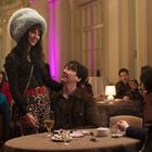 Eine Frau mit großem, grauem Hut steht lächelnd an einem Tisch in einem eleganten Restaurant. Mehrere Leute sitzen um sie.