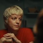 Eine Person mit kurzem, blondem Haar und rotem Pullover schaut zur Seite, Hände gefaltet vor sich. Hintergrund unscharf.