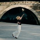 Eine Frau tanzt auf hellem Pflaster vor einer alten Steinbrücke in einer sonnigen Umgebung.
