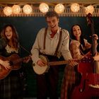 Drei Personen musizieren unter Lichtergirlanden. Eine Gitarre, ein Banjo und ein Kontrabass sind zu sehen. Alle lächeln.
