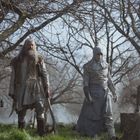 Zwei Figuren in altertümlicher Kleidung, eine mit langem Bart und Stock, stehen in einem nebligen Wald bei einem Grabstein.
