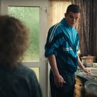 Ein Mann in einer blauen Trainingsjacke steht an einem Küchenspülbecken. Weitere Personen sitzen und stehen im Raum.