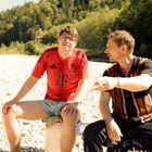 Ein Mann trägt ein rotes Fußballtrikot mit „T“ und Wappen. Sie sitzen auf einem steinigen Flussufer vor bewaldeter Landschaft.