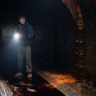 Keine sichtbaren Texte. Eine Person in einem dunklen Tunnel hält eine Taschenlampe. Leitern an der Wand.