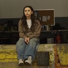 Eine junge Frau in Jeans und Jacke sitzt nachdenklich auf einer abgenutzten Mauer in einer Werkstatt.