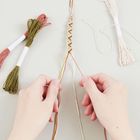 Zettel mit Texten: "EMBROIDERY FLOSS". Hände flechten ein mehrfarbiges Armband aus Fäden. Daneben liegen Fadenbündel.